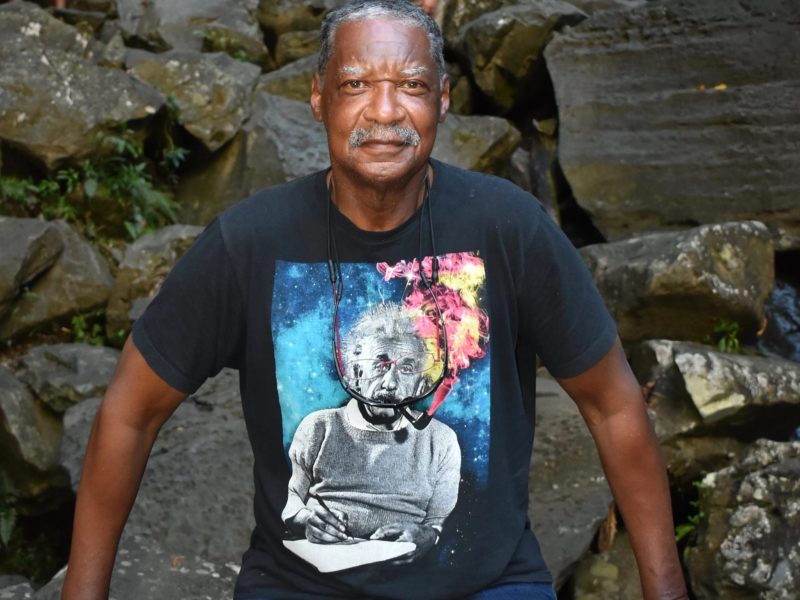 Eddie Conway, outside, in a t-shirt, standing in front of a waterfall