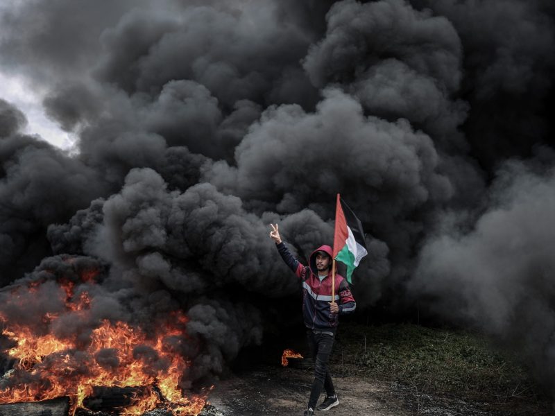 A young Palestinian man waves his flag in front of a thick column of black smoke