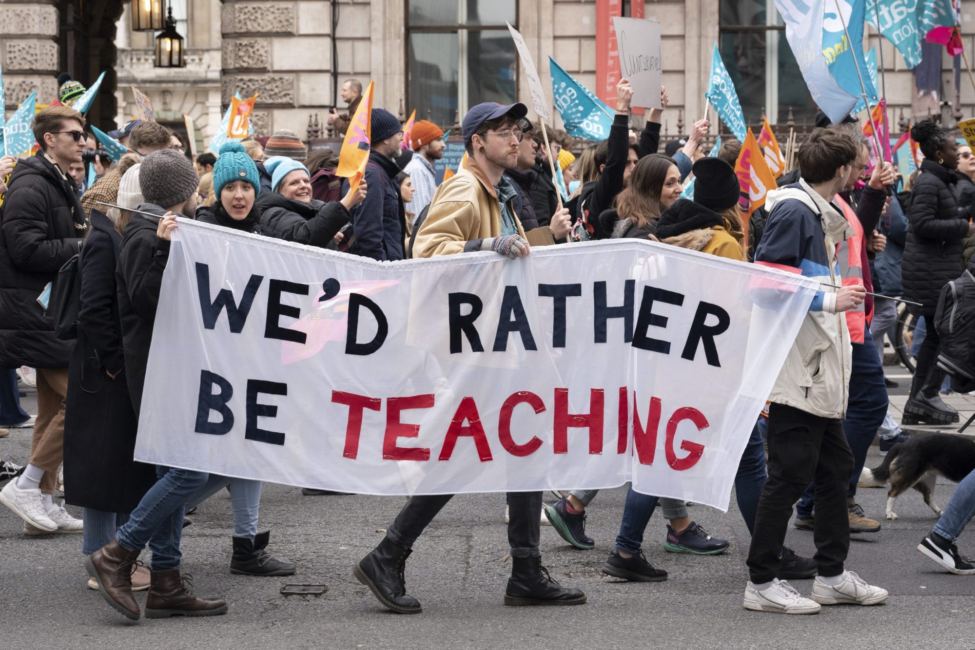 A group of striking teachers are seen marching in the street from the side. They are carrying a banner that says "We'd rather be teaching."