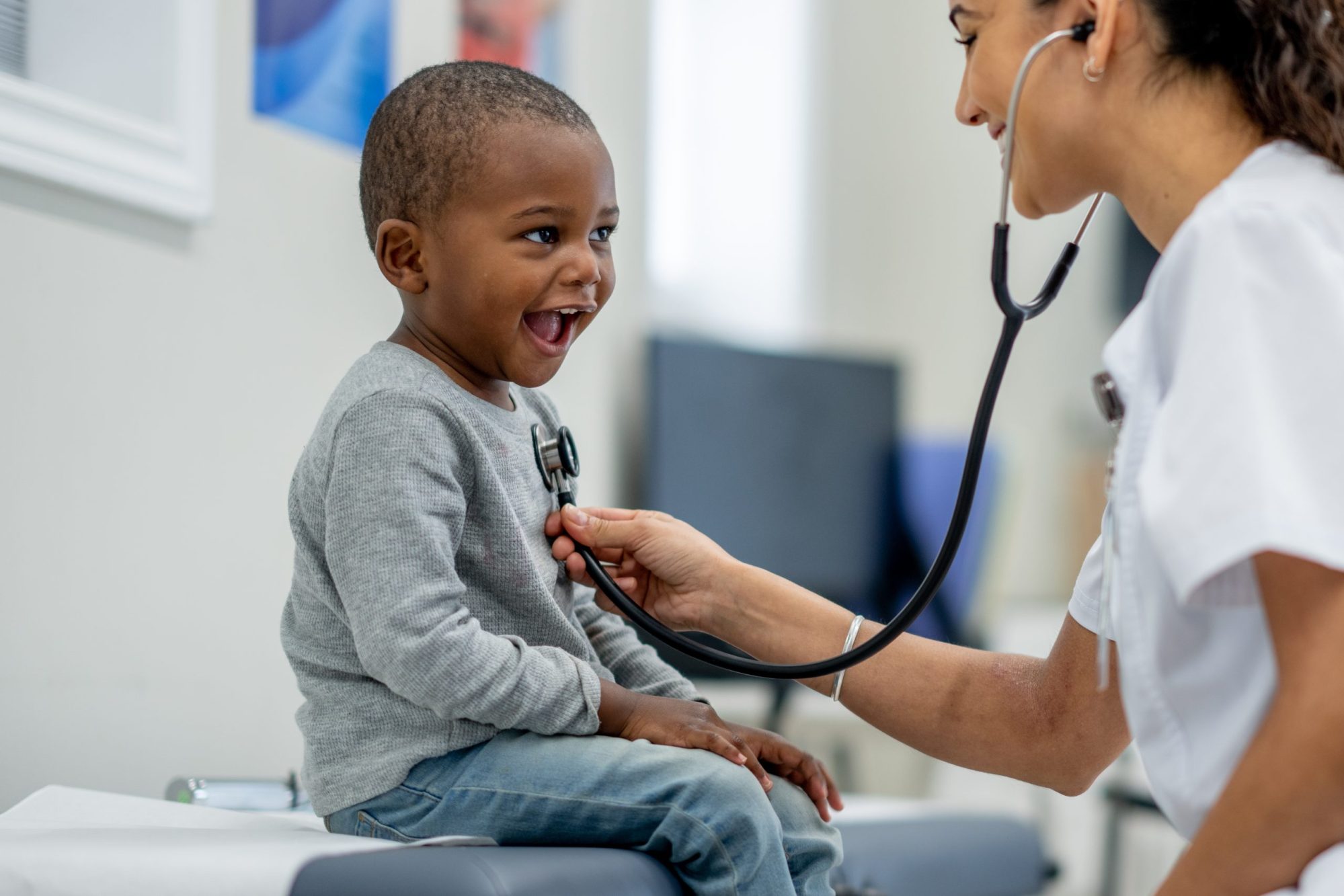 A young boy sits up on an exam table as a pediatrician preforms a check-up on him. Photo via Getty Images