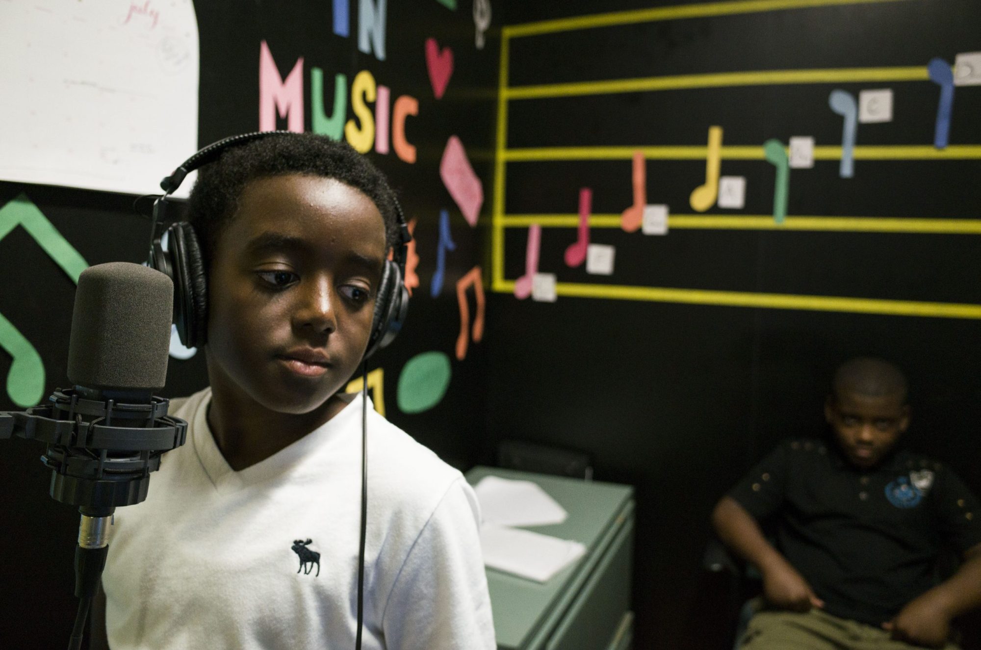 A young Black child wearing a white school uniform and a set of headphones stands in front of a microphone.