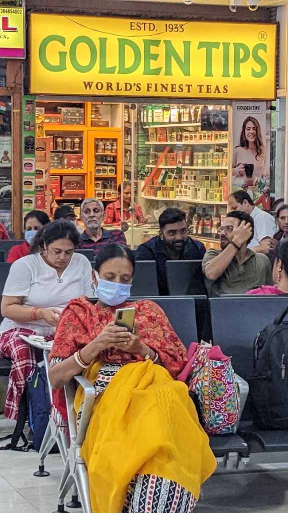 Travelers sit on benches talking, waiting, looking at their phones at the Bagdogra Airport in Bagdogra, Siliguri, West Bengal, India, on October 14, 2022. Behind them is a storefront with a big yellow sign that reads "ESTD. 1933, GOLDEN TIPS, WORLD'S FINEST TEAS."